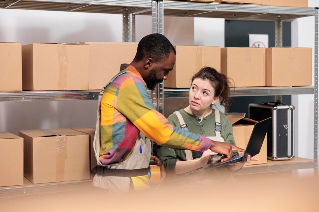 Deux collègues consultent un ordinateur portable devant des rayonnages de boîtes de carton pour gérer l'inventaire d'un local d'entreposage.